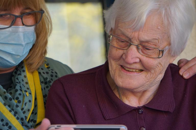 A woman in a face mask holds an elderly lady whilst she looks at a phone.